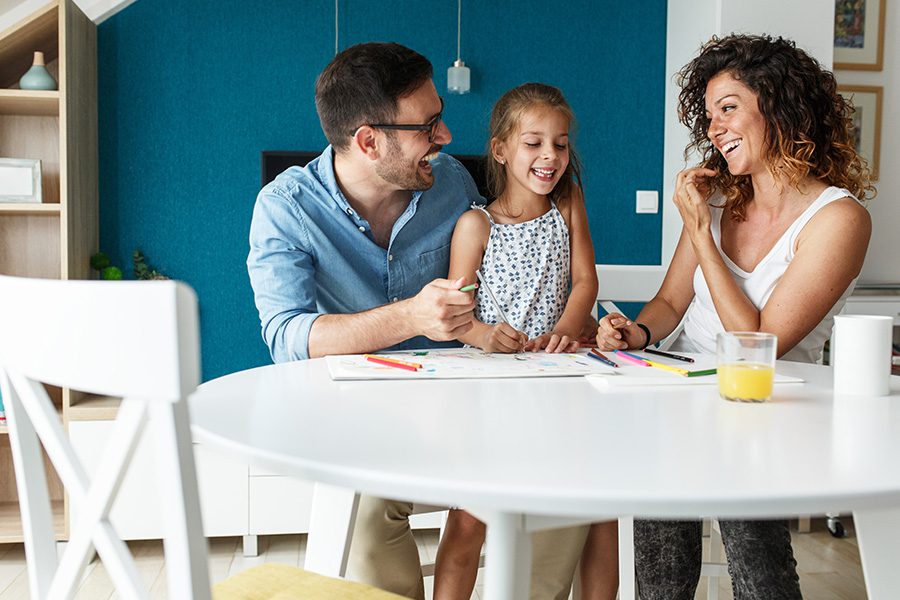 Personal Insurance - Father and Mother Smile and Laugh With Their Daughter as They All Sit Together at a Table to Draw